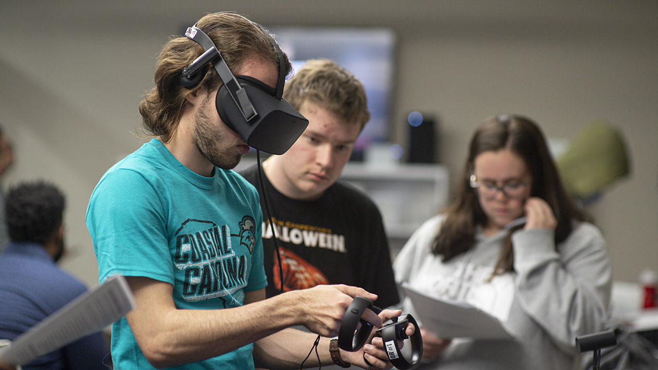 Students work with virtual reality equipment in a classroom, using handheld controllers while others review printed materials nearby.